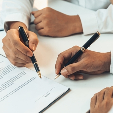 Two people's hands looking over a contract and holding pens.
