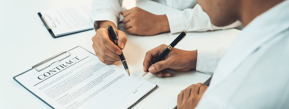 Two people's hands looking over a contract and holding pens.