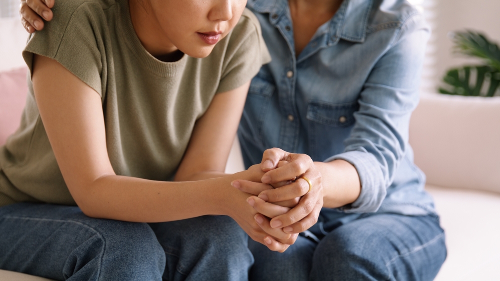 A worried woman seated on a sofa is comforted by another woman who clasps her hands.