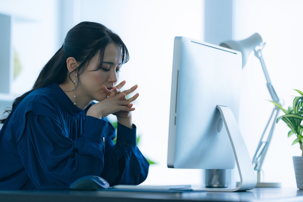 A woman seated in front of a computer with a worried mien and clasping her hands together.